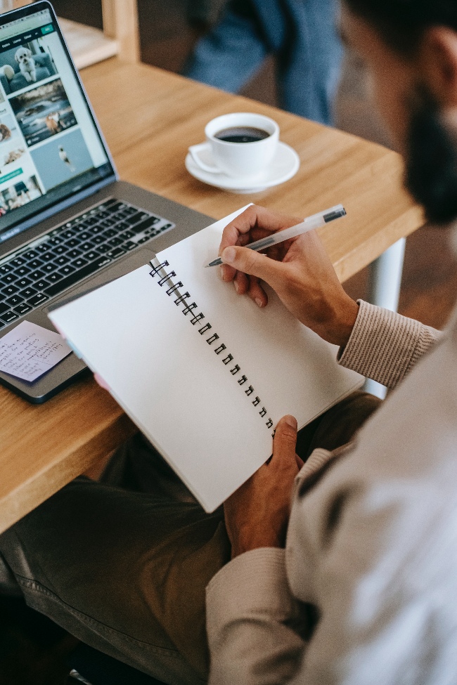 Partial view of a person sitting at a desk writing in a notepad in front of a laptop