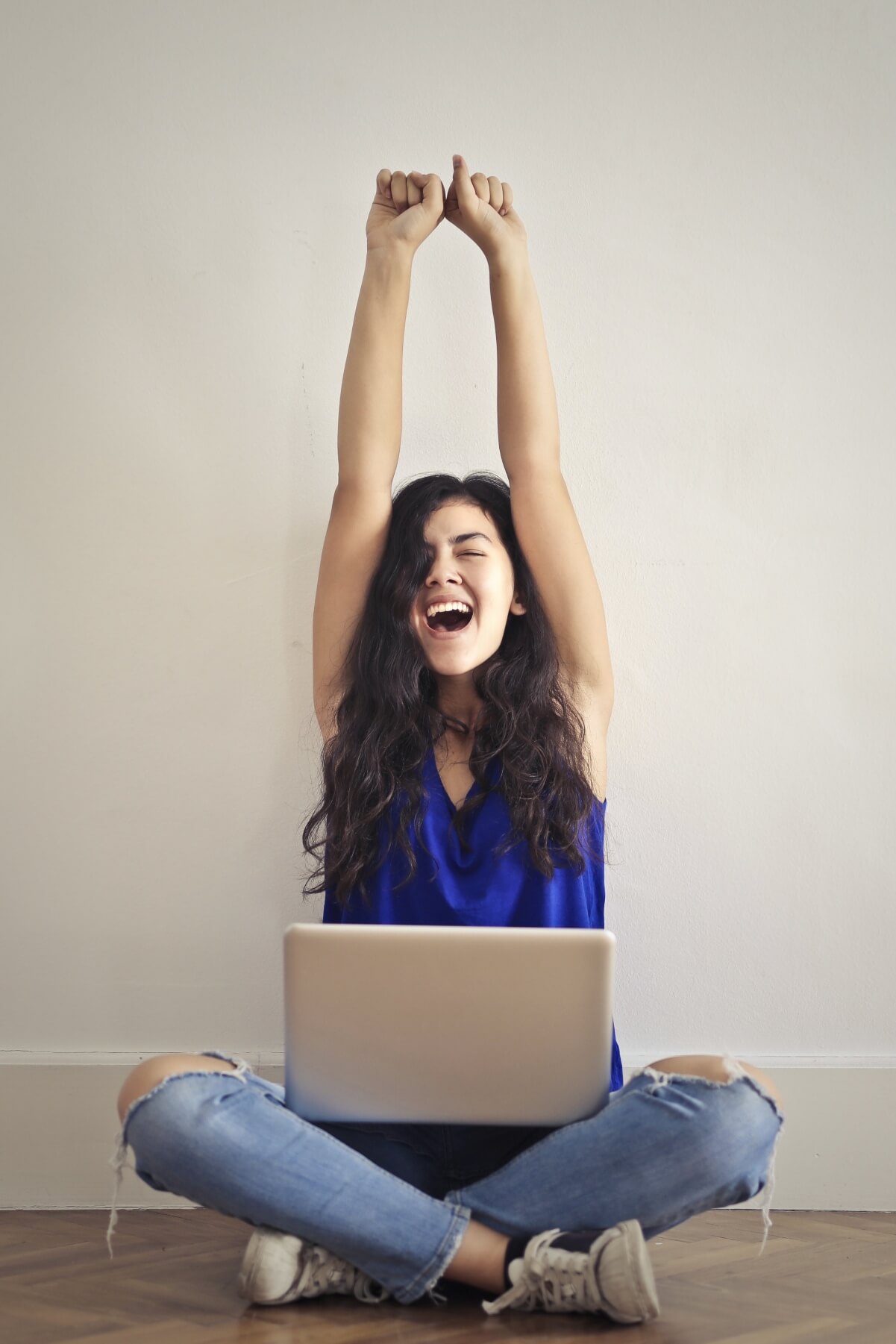 Young woman sitting on the floor with her laptop with her arms raised in excitement